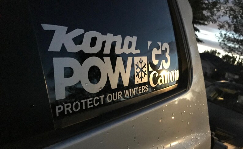May include: White sticker on a car window with the words "Kona POW" in large, stylized letters. Below, it reads "PROTECT OUR WINTERS". A snowflake graphic is included. The sticker is on a car.