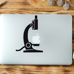 May include: A black microscope sticker with a white apple logo on a silver laptop. The laptop is on a wooden surface, with a blue notebook, pen, and white mouse nearby. Decorative lights and flowers are also present.