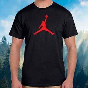 May include: Black t-shirt featuring a red silhouette of a person jumping with a basketball. The graphic is centered on the chest. The shirt has short sleeves and a crew neck. The background is a blurred image of a forest and sky.