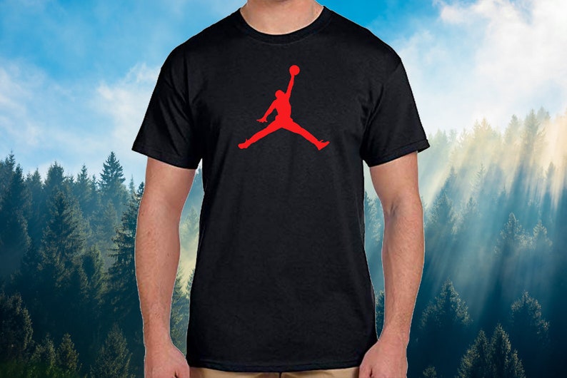 May include: Black t-shirt featuring a red silhouette of a person jumping with a basketball. The graphic is centered on the chest. The shirt has short sleeves and a crew neck. The background is a blurred image of a forest and sky.