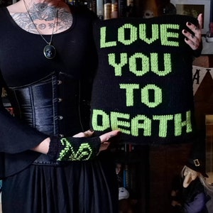 以下が含まれることがあります： 緑色の糸で「LOVE YOU TO DEATH」と刺繍された黒いニットのクッション