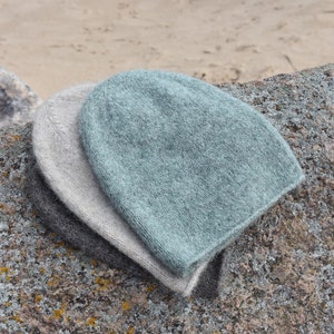 May include: Two knitted hats, one light grey and one light blue, are stacked on top of each other on a grey rock.