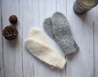 Könnte beinhalten: Ein Paar Fäustlinge, einer in Creme und einer in meliertem Grau, beide mit einer strukturierten, flauschigen Strickoptik und gerippten Bündchen. Die Fäustlinge sind zusammen mit zwei kleinen braunen Tannenzapfen und einer Kerze in einem dunklen Glasgefäß abgebildet.