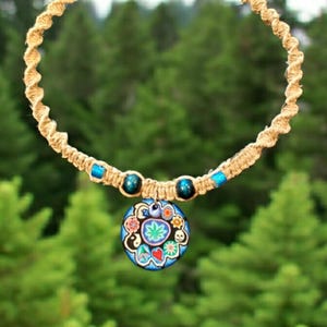 May include: A hemp necklace with a blue and green pendant featuring a variety of symbols, including a peace sign, a heart, and a skull. The necklace is made with natural hemp cord and blue beads.