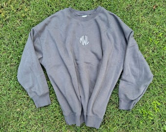 May include: A charcoal grey crewneck sweatshirt featuring long sleeves with ribbed cuffs and a ribbed hem. The centre of the chest displays the embroidered letters MWL in a light grey, cursive-style font, creating a simple and clean design.