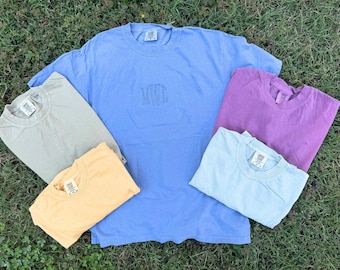 May include: A collection of five short-sleeved crew neck t-shirts in various solid colours including periwinkle blue, beige, light orange, purple, and light blue. The central periwinkle shirt features the embroidered monogram letters MWL.