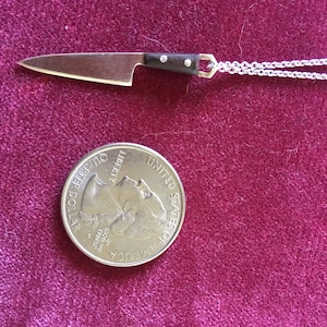 May include: A silver chain necklace with a miniature knife pendant. The knife has a silver blade and a black wooden handle. The pendant is about the size of a 20 pence coin.