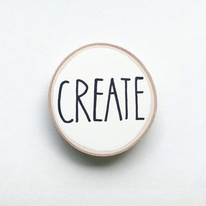 May include: A round, wooden embroidery hoop with the word "CREATE" in black, handwritten-style lettering centered on a white background. The hoop has a natural wood finish. The background is plain white.