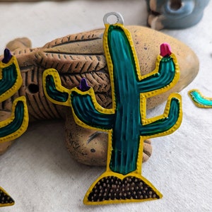 May include: A green and yellow metal cactus ornament with pink and purple accents. The cactus is standing on a brown base with a textured surface.