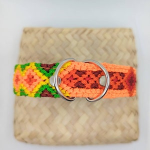 Puede incluir: Un collar para mascotas de tela tejida con un patrón geométrico vibrante en tonos naranja, amarillo, verde y marrón oscuro. El collar incluye dos anillos en D de metal plateado asegurados con remaches metálicos para mayor resistencia.