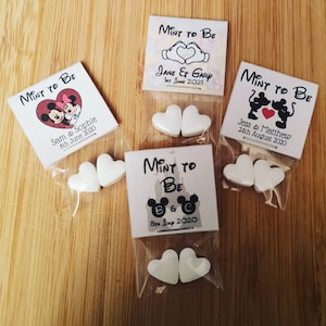 Könnte beinhalten: Vier weiße Beutel mit durchsichtigen Cellophanfenstern, die jeweils zwei weiße herzförmige Minzen enthalten. Jeder Beutel hat eine weiße quadratische Karte mit einem Disney-Motiv und dem Text "Mint to Be" sowie den Namen eines Paares und das Hochzeitsdatum.