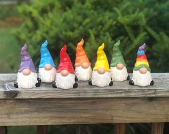May include: Seven colourful ceramic gnomes with white beards and different coloured hats. The gnomes are arranged in a row on a wooden surface.