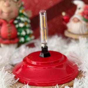 Peut inclure: Un support de sapin de Noël en métal rouge avec une base noire et une ampoule transparente. Le support est posé sur une surface blanche et moelleuse.