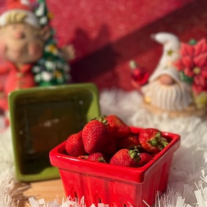 May include: A red ceramic basket filled with fresh red strawberries. The basket has a lattice design and is sitting on a white fluffy surface.