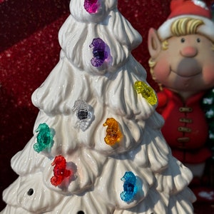 May include: A white ceramic Christmas tree with colorful seahorse ornaments. The ornaments are purple, blue, green, yellow, red, and clear. The tree is decorated with small holes.