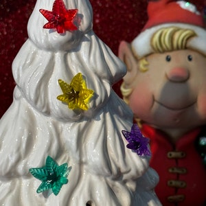 May include: A white ceramic Christmas tree with four colorful light bulbs. The bulbs are red, yellow, green, and purple. The tree is decorated with a white, fluffy texture. A Christmas elf is standing behind the tree.