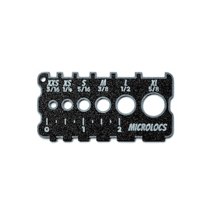 May include: A black rectangular Microlocs sizing tool featuring six circular cutouts labeled XXS 3/16, XS 1/4, S 5/16, M 3/8, L 1/2, and XL 5/8. The tool includes a printed ruler along the bottom edge marked from 0 to 2 inches for precise measurements.