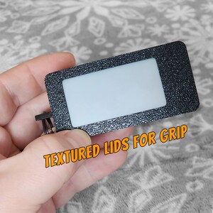 Pode incluir: Uma etiqueta retangular preta com uma janela transparente e uma superf&iacute;cie texturizada. A etiqueta tem um anel de metal preso a ela. O texto "TEXTURED LIDS FOR GRIP" est&aacute; impresso na etiqueta.