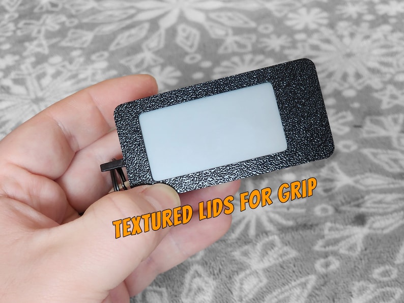 Pode incluir: Uma etiqueta retangular preta com uma janela transparente e uma superf&iacute;cie texturizada. A etiqueta tem um anel de metal preso a ela. O texto "TEXTURED LIDS FOR GRIP" est&aacute; impresso na etiqueta.