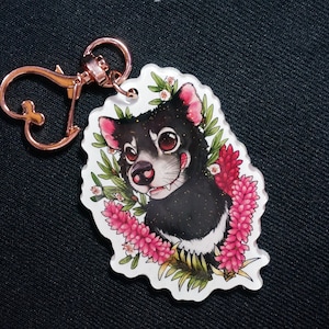 May include: A black and white Tasmanian devil cartoon character with pink flowers and leaves around it. The character is on a clear acrylic keychain with a rose gold metal loop.