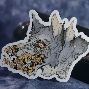 May include: A sticker featuring a detailed illustration of a wolf's head with a snarling expression. The wolf's fur is a light gray color, and its eyes are a deep blue. The wolf's mouth is open, revealing sharp teeth and a tongue. The wolf's head is surrounded by a scattering of pink flowers.