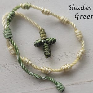 May include: A green and cream knotted cord bracelet with a small green cross charm. The bracelet has the text "Shades of Green" written on the image.