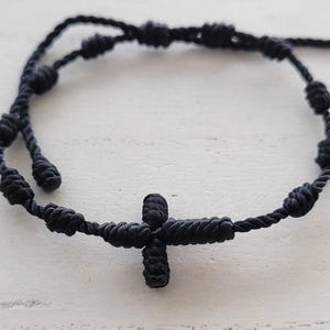 May include: A black knotted cord bracelet featuring a cross pendant. The bracelet is made of woven black cord with several knots along the length. The cross is also made of knotted cord.