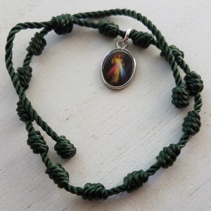 May include: A green knotted cord bracelet with a silver charm featuring an image of Jesus. The charm is oval shaped and has a white background with a colorful image of Jesus.