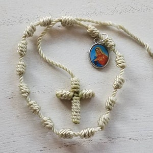 Puede incluir: Un rosario de cuerda anudada de color verde claro. El rosario tiene una cruz pequeña y un colgante con una imagen de la Virgen María.