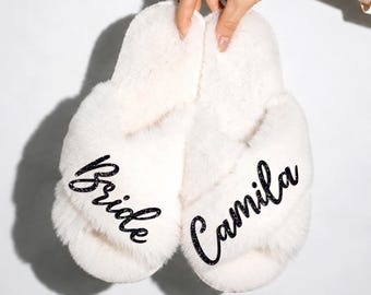 May include: A pair of white plush faux fur slippers with a crisscross strap design. The left slipper features the word Bride in black script, and the right slipper features the name Camila in black script, both written in a glittery finish.