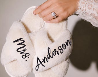 May include: A pair of cream-colored faux fur slippers with a crisscross strap design. The left slipper features the black glittery text Mrs. and the right slipper features the black glittery text Anderson printed on the top of the plush fabric straps.