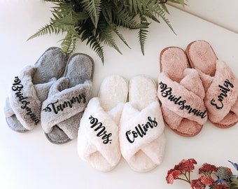 May include: Three pairs of plush, open-toe slippers with crisscross straps. The gray pair features the text "Bridesmaid" and "Tammy," the white pair features "Mrs." and "Collins," and the pink pair features "Bridesmaid" and "Emily" in black script.