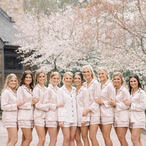 May include: A group of ten individuals wearing matching light pink button-down pajama sets consisting of long-sleeved shirts and shorts with dark piping detail along the collars, cuffs, and hems, standing together in a row outdoors.
