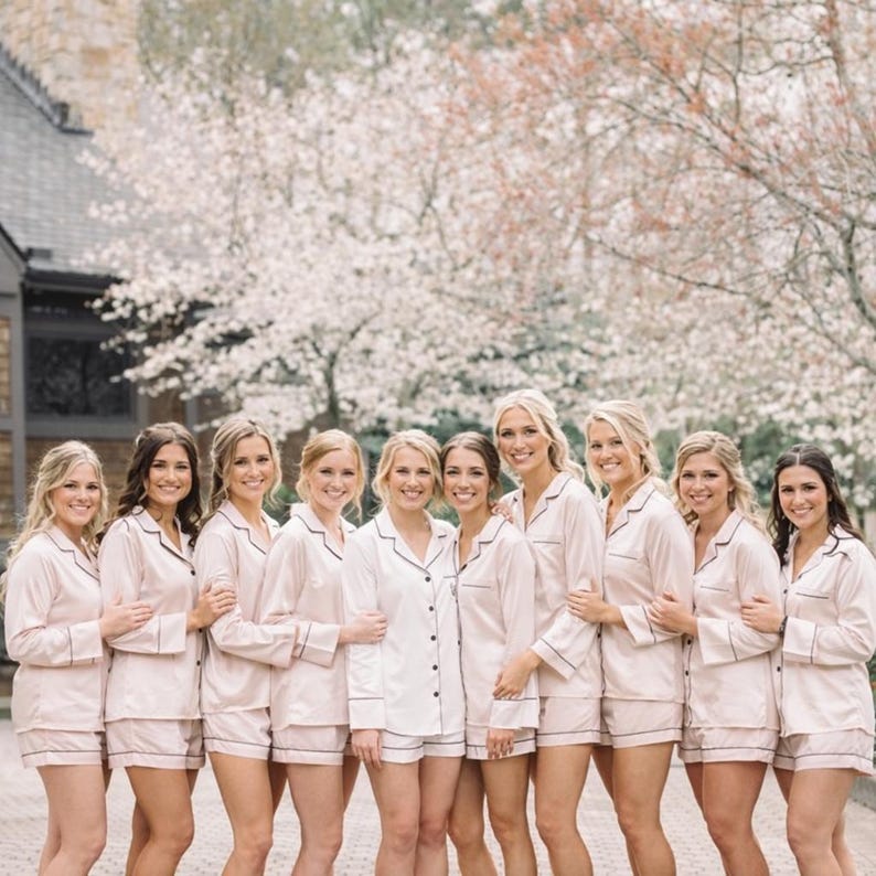 May include: A group of ten individuals wearing matching light pink button-down pajama sets consisting of long-sleeved shirts and shorts with dark piping detail along the collars, cuffs, and hems, standing together in a row outdoors.