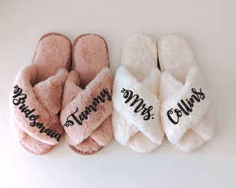 May include: Two pairs of plush, open-toe slippers with crisscross straps. One pair is dusty rose with black glitter text reading Bridesmaid and Tammy. The other pair is white with black glitter text reading Mrs. and Collins on the straps.