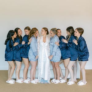 May include: A group of people wearing matching navy blue and light blue pajama sets consisting of button-down shirts and shorts with white piping, paired with white fuzzy slippers. One person wears a long white lace robe over a white slip dress.
