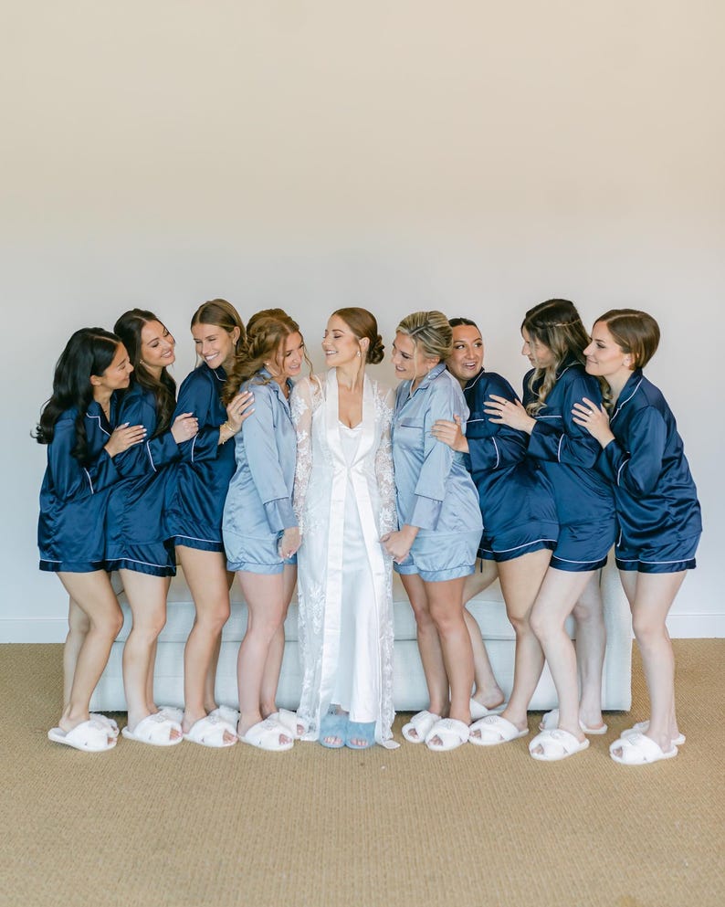 May include: A group of people wearing matching navy blue and light blue pajama sets consisting of button-down shirts and shorts with white piping, paired with white fuzzy slippers. One person wears a long white lace robe over a white slip dress.