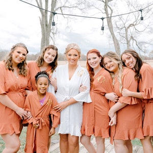 May include: A group of women and a young girl wearing matching rust-colored robes with ruffled sleeves and gold lettering. The women are smiling and posing for a photo. The bride is in the center wearing a white robe.