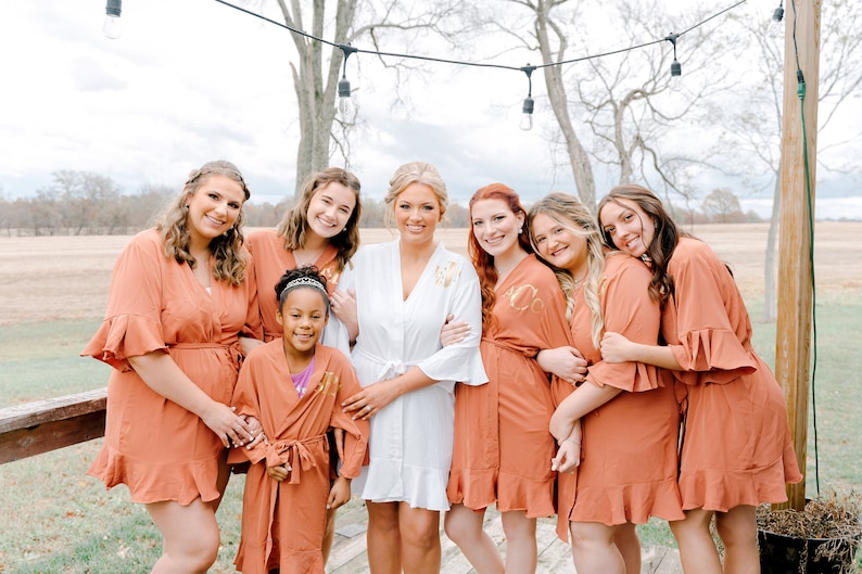 May include: A group of women and a young girl wearing matching rust-colored robes with ruffled sleeves and gold lettering. The women are smiling and posing for a photo. The bride is in the center wearing a white robe.