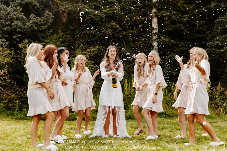 May include: A group of women wearing white and cream-colored robes are celebrating with champagne. They are standing in a grassy area with trees in the background. The text "BRIDESMAID ROBES BY SWEET NOVEMBER GIFTS OUR HAPPY CUSTOMERS WERE WEARING OAT COLOR" is visible at the bottom of the image.