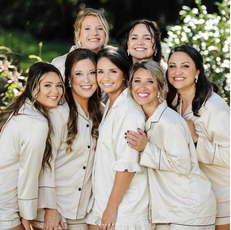 May include: A group of six individuals wearing matching champagne-colored button-down pajama sets. The shirts feature long sleeves, collars, and dark piping details along the edges. The fabric has a smooth, satin-like finish and a light sheen.
