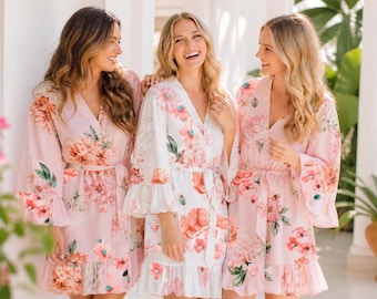 May include: Three robes featuring a floral pattern with pink, peach, and green blossoms. Two robes are light pink, and one is white. Each robe has a V-neckline, long flared sleeves, a matching fabric tie belt at the waist, and a ruffled hemline.