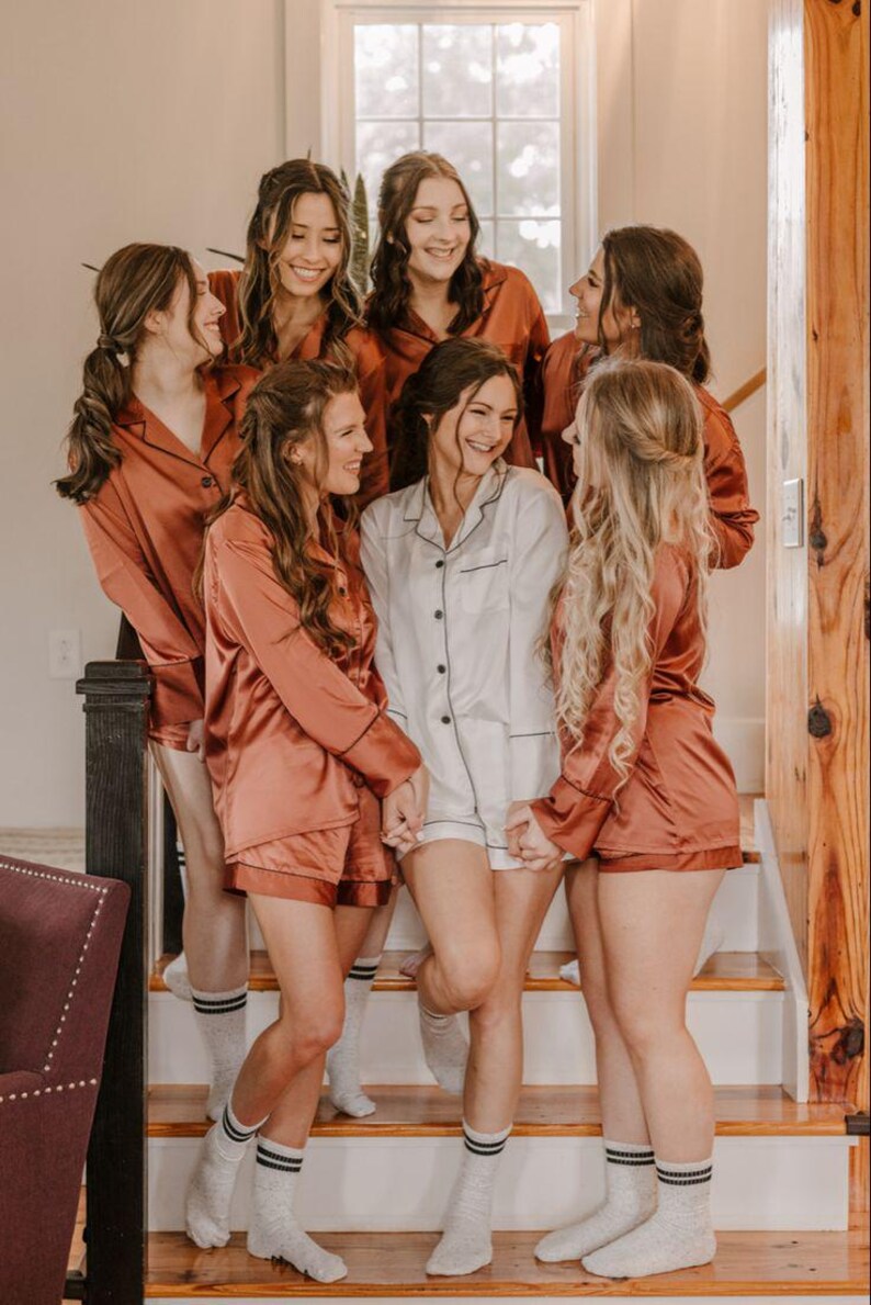 May include: A group of individuals wearing matching button-down pajama sets with long sleeves and shorts. One individual wears a white set with black piping, while the others wear rust-colored sets with black piping. All are wearing white crew socks.