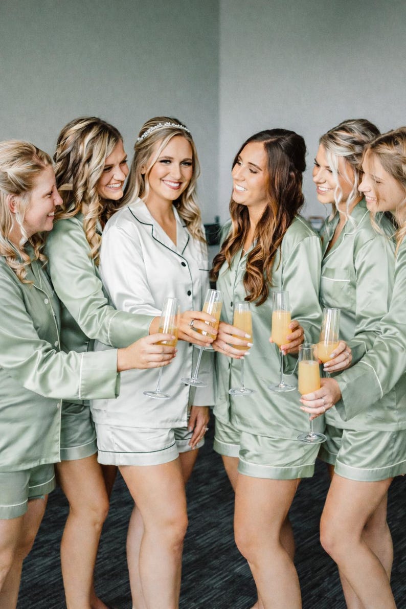 May include: A group of six individuals wearing matching button-down pajama sets in sage green and white satin fabric with contrasting piping. Each person holds a tall, slender glass filled with a yellow liquid, celebrating together in coordinated sleepwear.