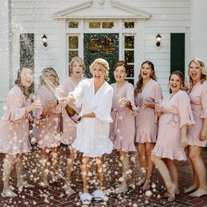 May include: A group of women in pink robes celebrate with champagne. The bride in a white robe pops the bottle, spraying the group with bubbly.