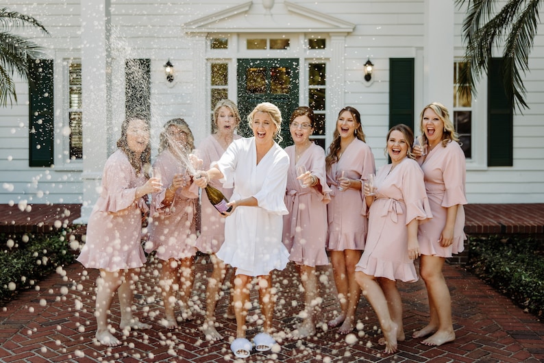 May include: A group of women in pink robes celebrate with champagne. The bride in a white robe pops the bottle, spraying the group with bubbly.