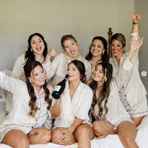 May include: A group of six people wearing matching champagne-colored satin pajama sets with dark piping. The sets consist of button-down shirts with collars and short bottoms. One person in the center holds a dark glass bottle of sparkling wine.