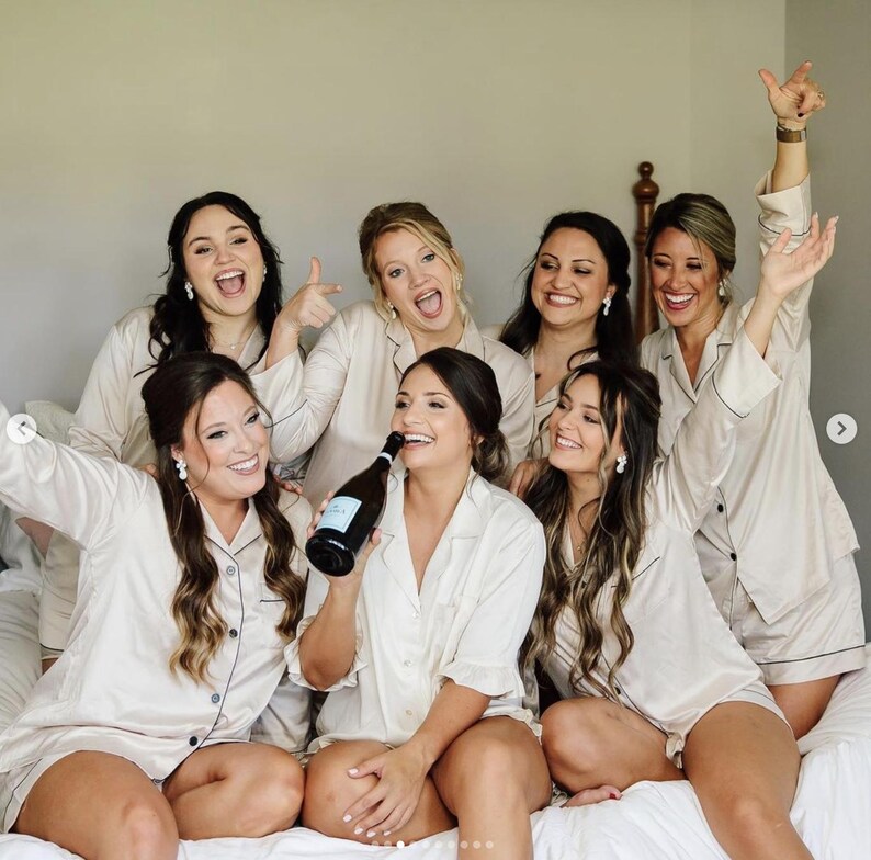 May include: A group of six people wearing matching champagne-colored satin pajama sets with dark piping. The sets consist of button-down shirts with collars and short bottoms. One person in the center holds a dark glass bottle of sparkling wine.