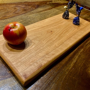 以下が含まれることがあります： 丸みを帯びた縁のある長方形の木製のまな板で、明るい茶色のチェリーウッドで作られています。赤いリンゴがボードの左側に置かれ、青と金のラッピングされたキャンディーが右側に置かれています。