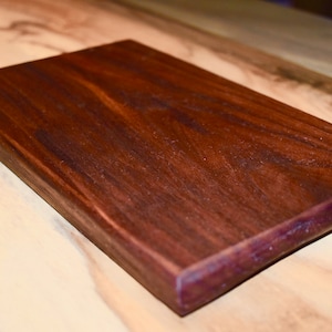 以下が含まれることがあります： 濃い茶色の木製のまな板で、生きたエッジと紫がかった色合いがあります。まな板は一枚の木材で作られており、滑らかで磨かれた仕上げになっています。
